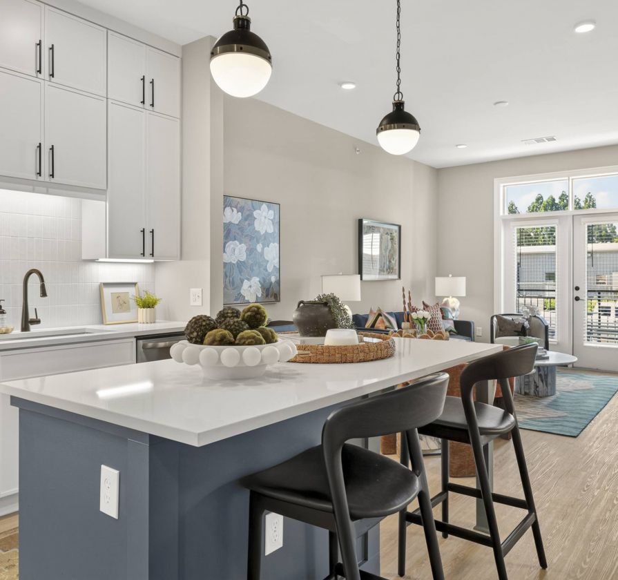 Modern kitchen with white cabinets, pendant lights, a large island, and black barstools, open to a bright living area with large windows.
