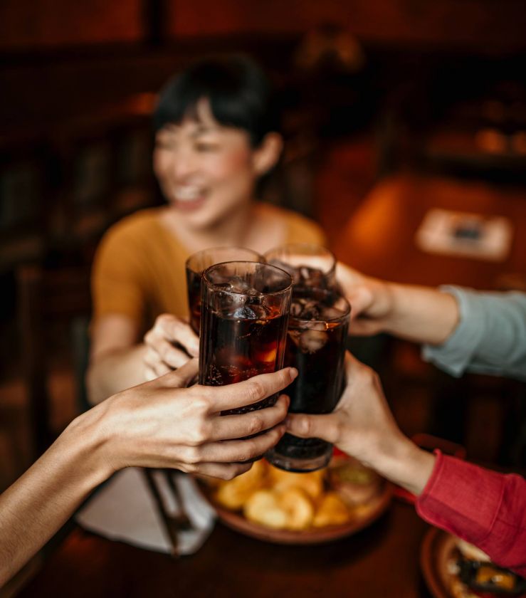 Three people clink glasses of soda together at a table, smiling, with snacks in the background.
