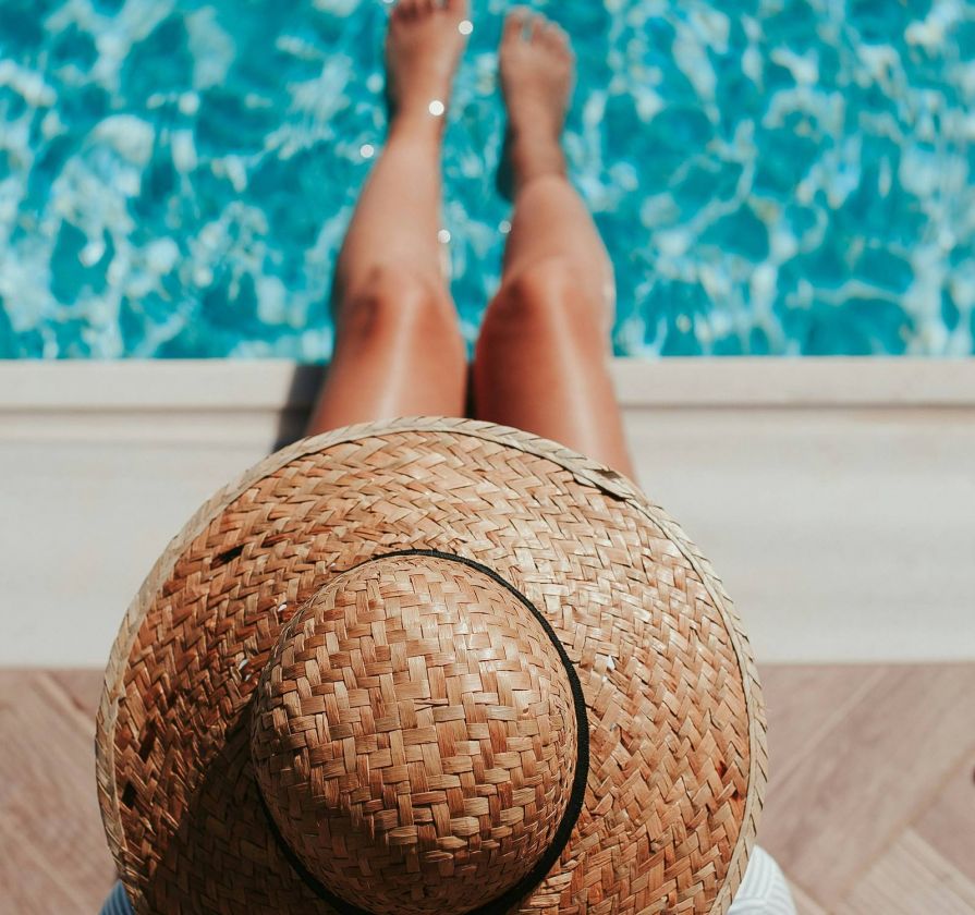 Person wearing a straw hat sits by a swimming pool with their legs stretched out toward the water.