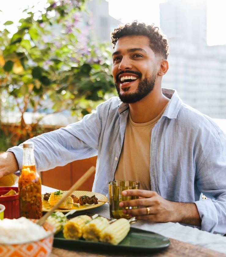 Man smiling at an outdoor table, holding a drink, surrounded by food and greenery with a cityscape in the background.