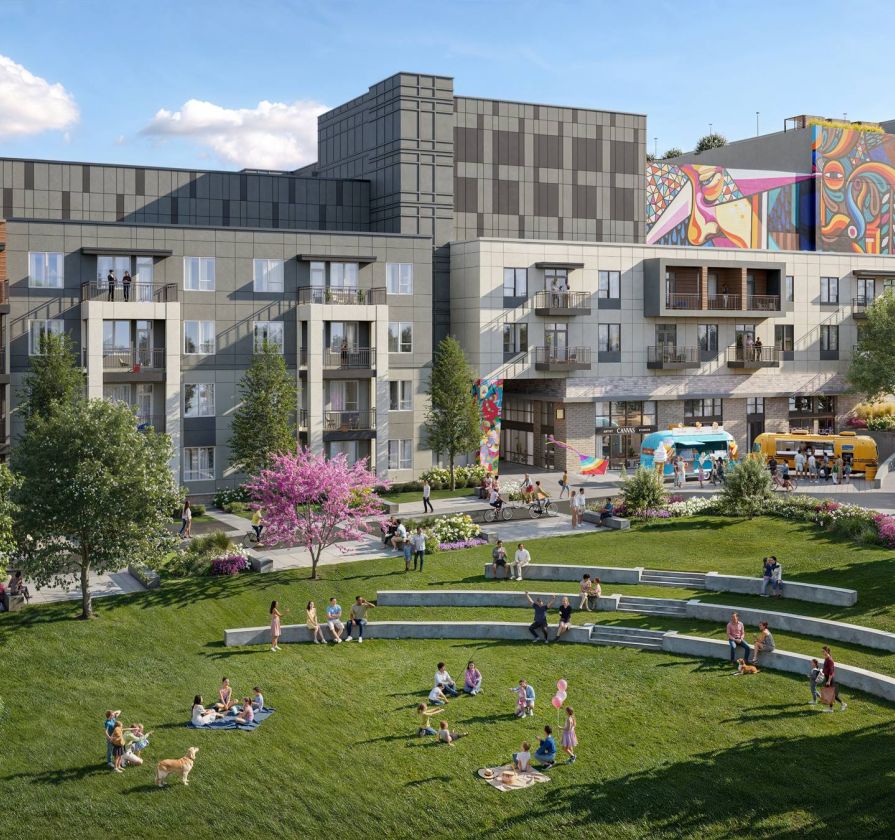 People relax on a grassy lawn in front of modern apartments with murals and outdoor seating on a sunny day.