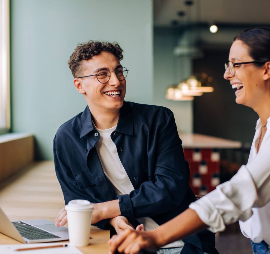 Two people sit at a table with a laptop and coffee, smiling and talking in a brightly lit, modern space.