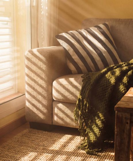 Sunlight streams through blinds onto a cozy sofa with striped pillow and green blanket beside a rustic coffee table.