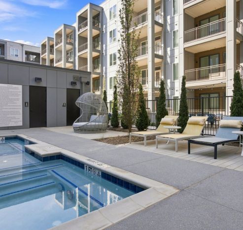 Three people enjoy a small outdoor pool and hot tub in a modern apartment complex courtyard on a sunny day.
