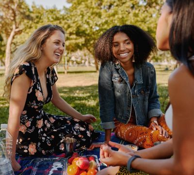 Three women sitting on a blanket in a park, smiling and talking on a sunny day.