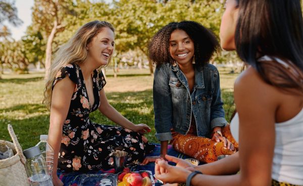 Three women sitting on a blanket in a park, smiling and talking on a sunny day.