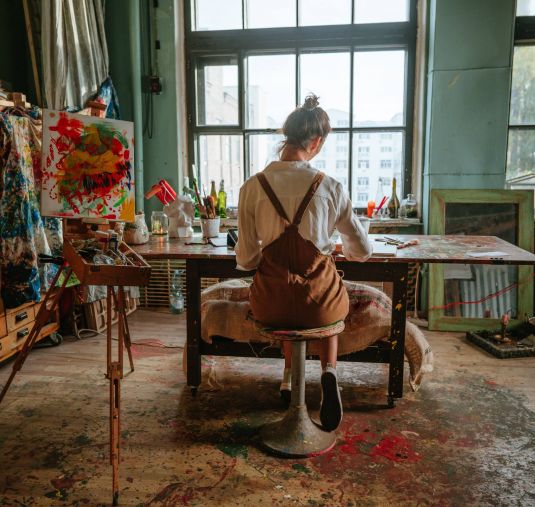 An artist sits at a wooden table in a sunlit studio, painting with supplies scattered around.