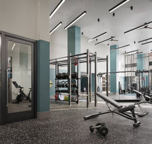 Modern gym with weight racks, benches, exercise bike, mirrored walls, and overhead lighting in a spacious, clean room.