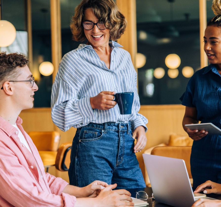 Three people having a casual meeting in a modern office, with laptops and coffee, smiling and talking together.
