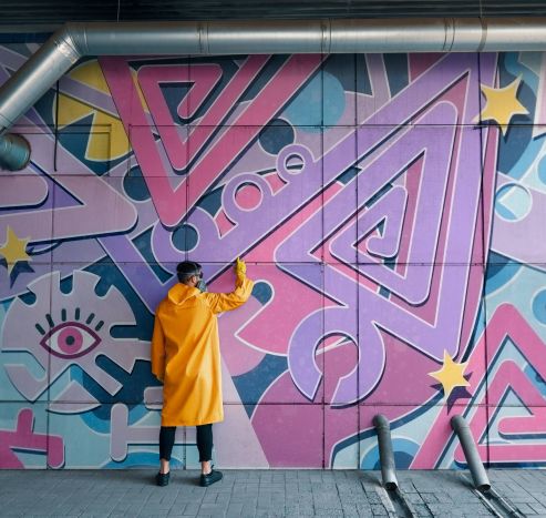 Person in yellow coat paints colorful geometric mural with pink, purple, and blue shapes on a wall.
