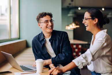 Two people sit at a table with a laptop and coffee, smiling and talking in a brightly lit, modern space.