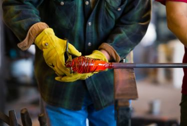 A person in yellow gloves shapes molten glass on a rod, wearing a green plaid shirt and blue jeans.