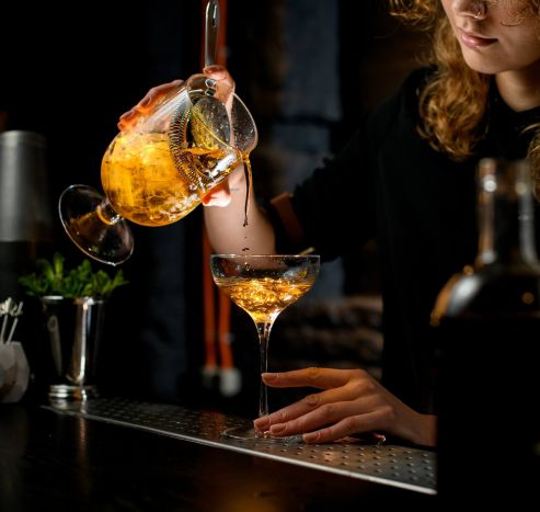 Bartender pouring an amber cocktail from a mixing glass into a coupe glass at a dimly lit bar.