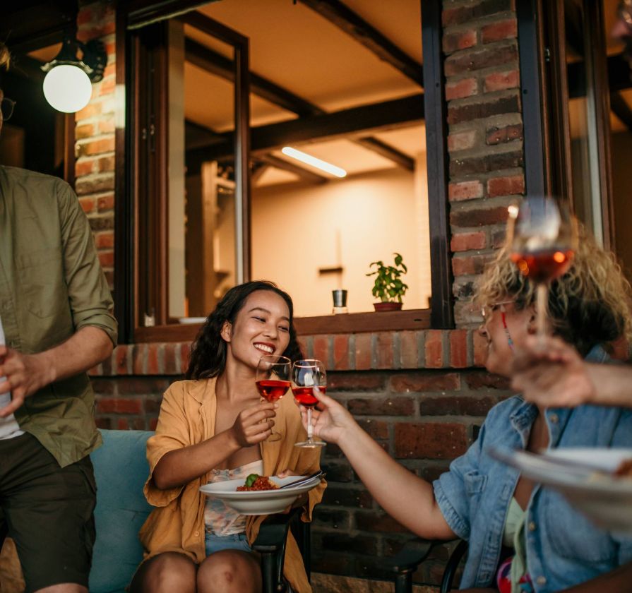 Three friends sit outside, smiling and clinking glasses of wine near a brick wall and open window.